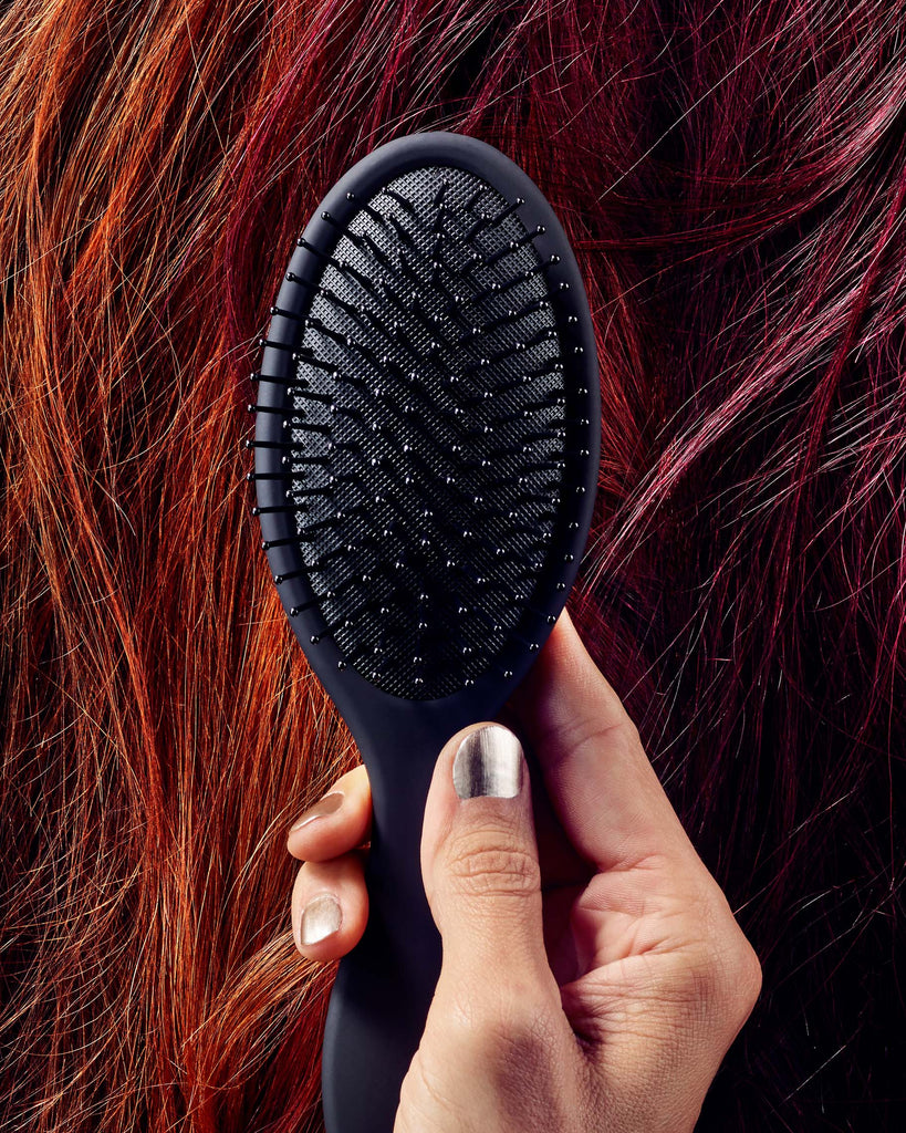 Close up of Everything Hand Tied's Cushion Paddle Brush held in front of various colors of Hand Tied Hair Extensions